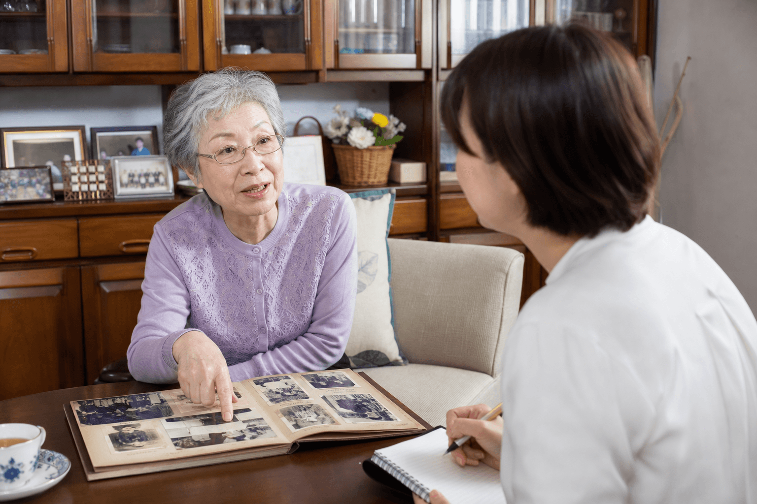 お客様がご自宅のリビングで、昔のアルバムを指差しながらライターに思い出を語っている取材風景の写真]